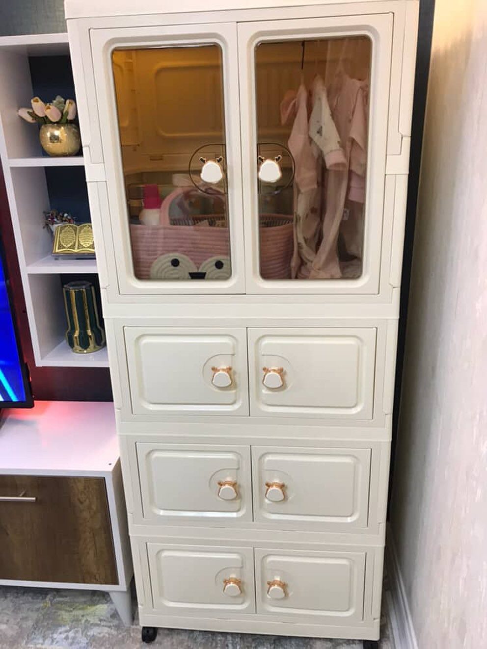 White baby wardrobe with glass doors containing pink clothes and storage baskets.