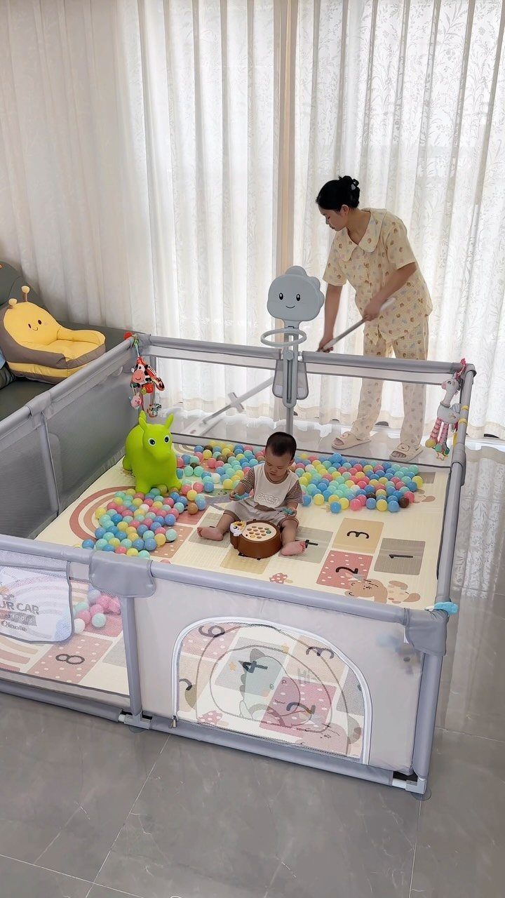 Baby and woman in a grey playpen with colorful balls and play mat.