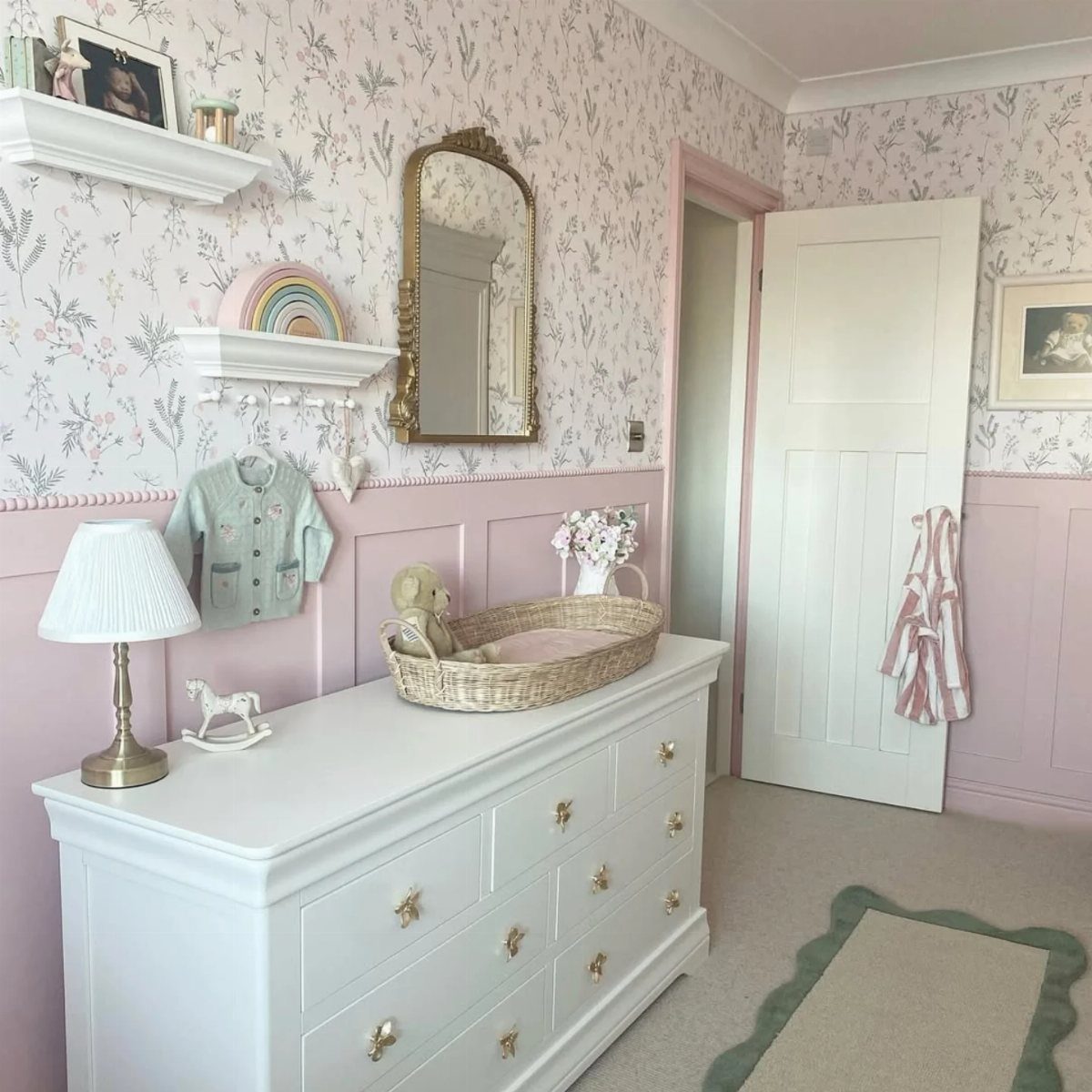 Nursery room with pink wainscoting, floral wallpaper, white dresser, and green-edged rug.