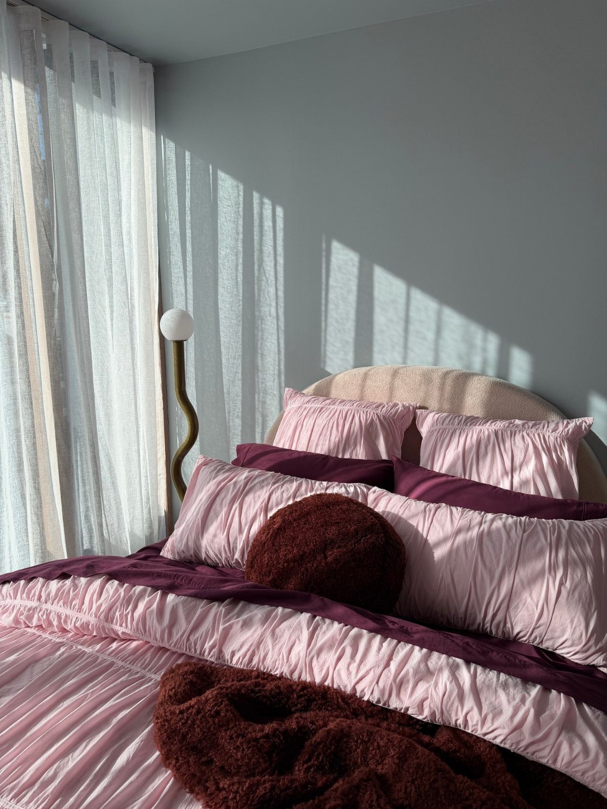 Bed with light pink pleated bedding and deep plum blankets against grey walls.