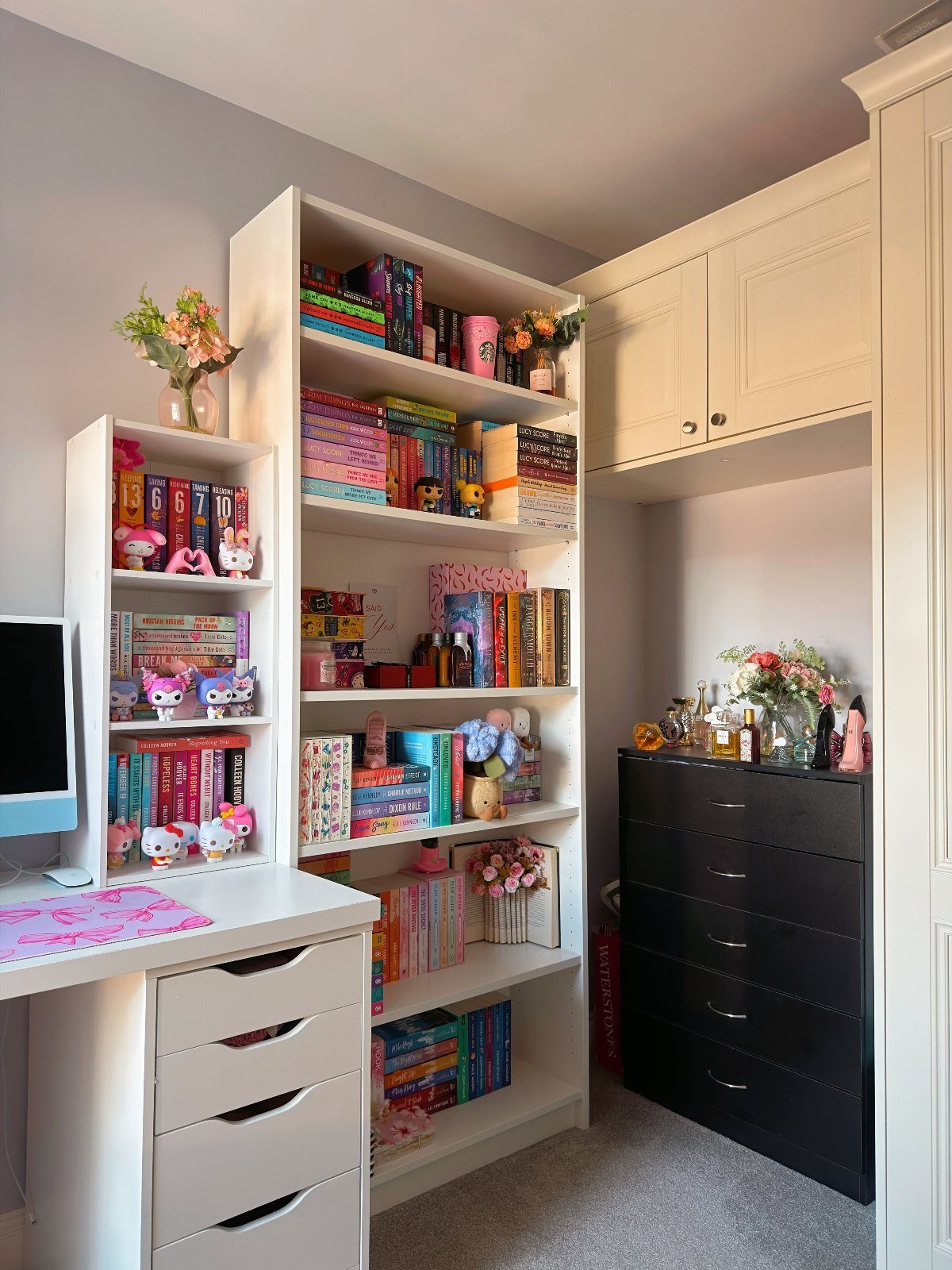 White tall bookshelf filled with colorful books and figurines next to a white desk.