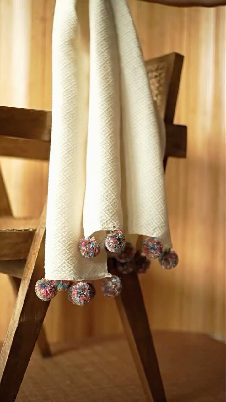 Cream textured throw blanket with colorful pom-poms draped over a wooden chair.