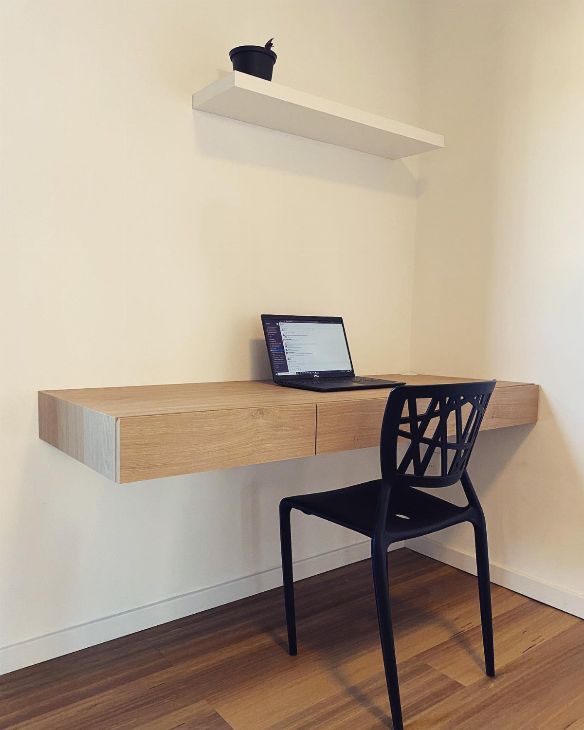 Light wood floating desk with a black chair and laptop against white walls.
