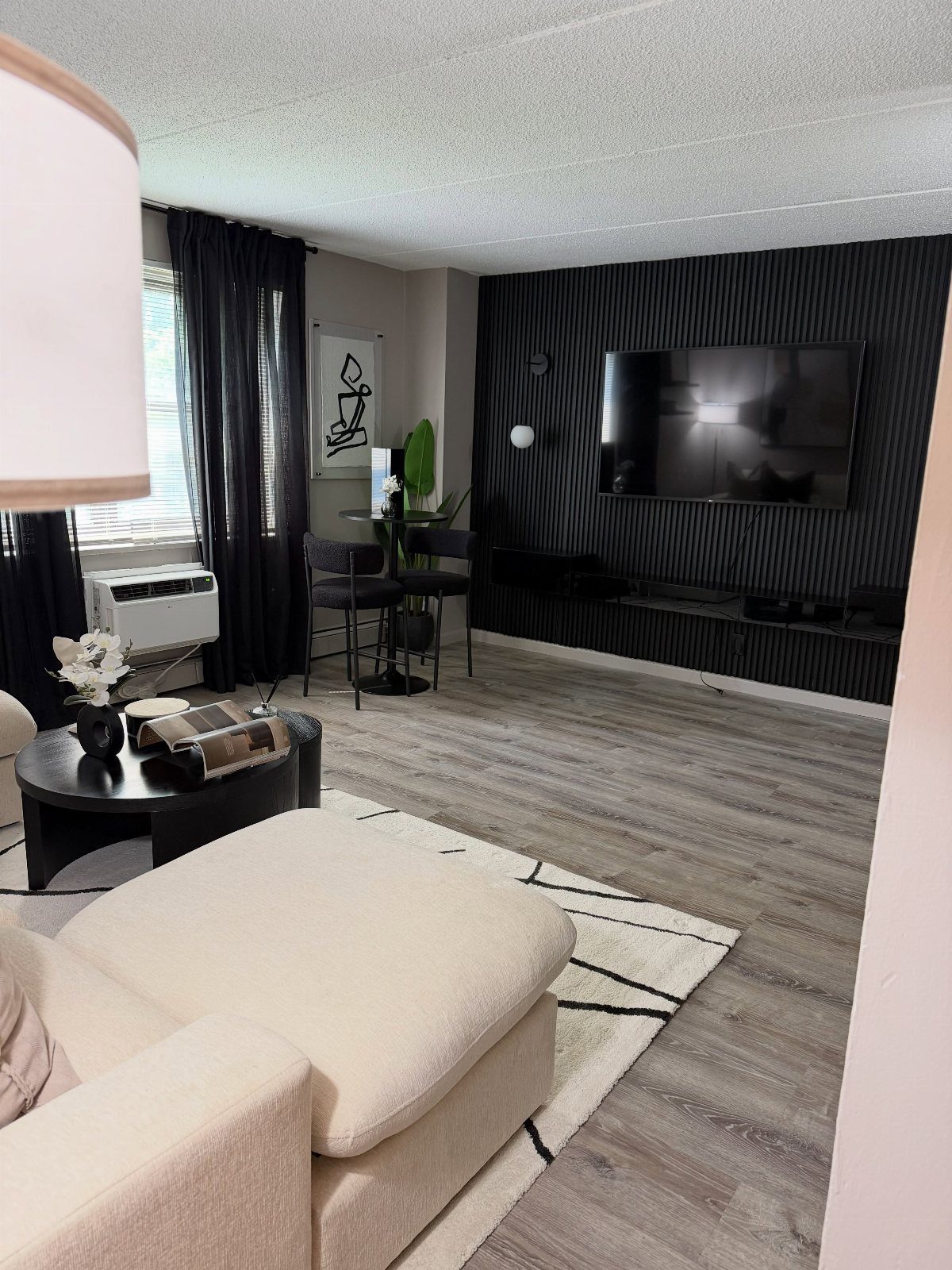 Living room with a black accent wall, white ottoman, and grey wood flooring.
