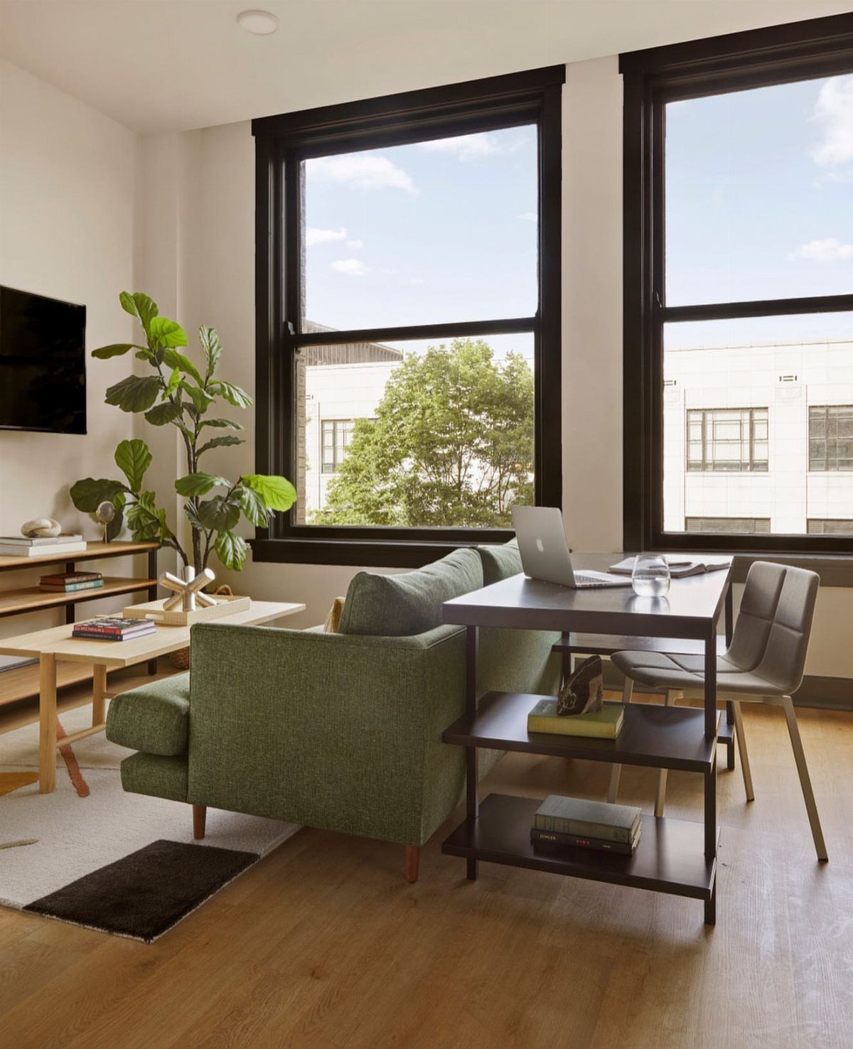 Living room with green sofa and integrated workspace by large black-framed windows.