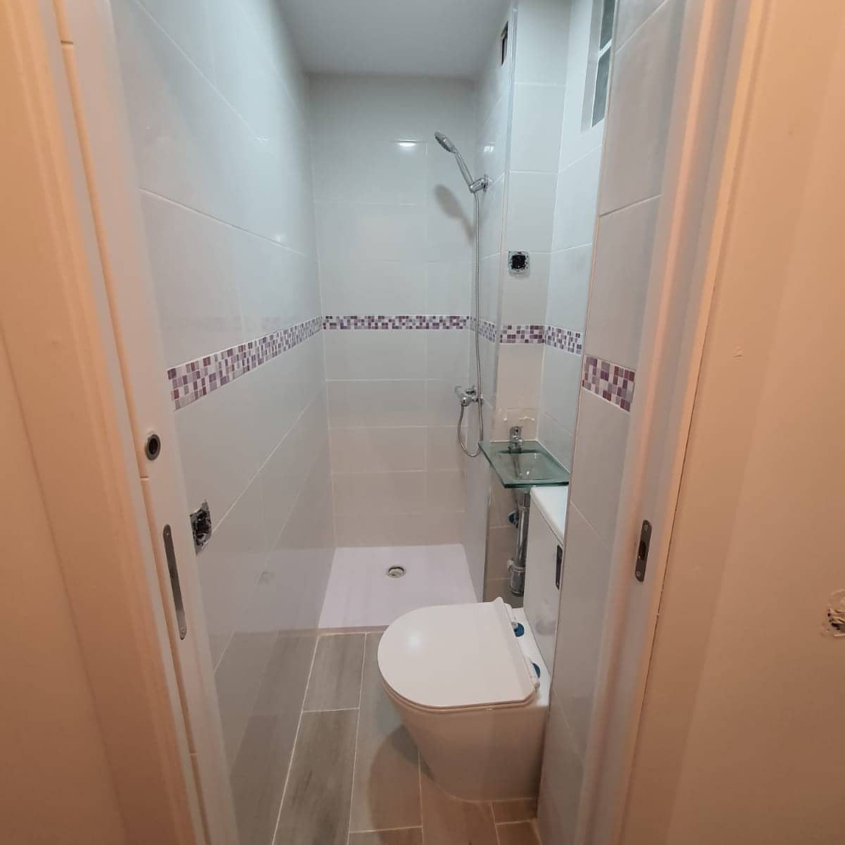 Narrow white tiled bathroom with a toilet and shower seen through open doors