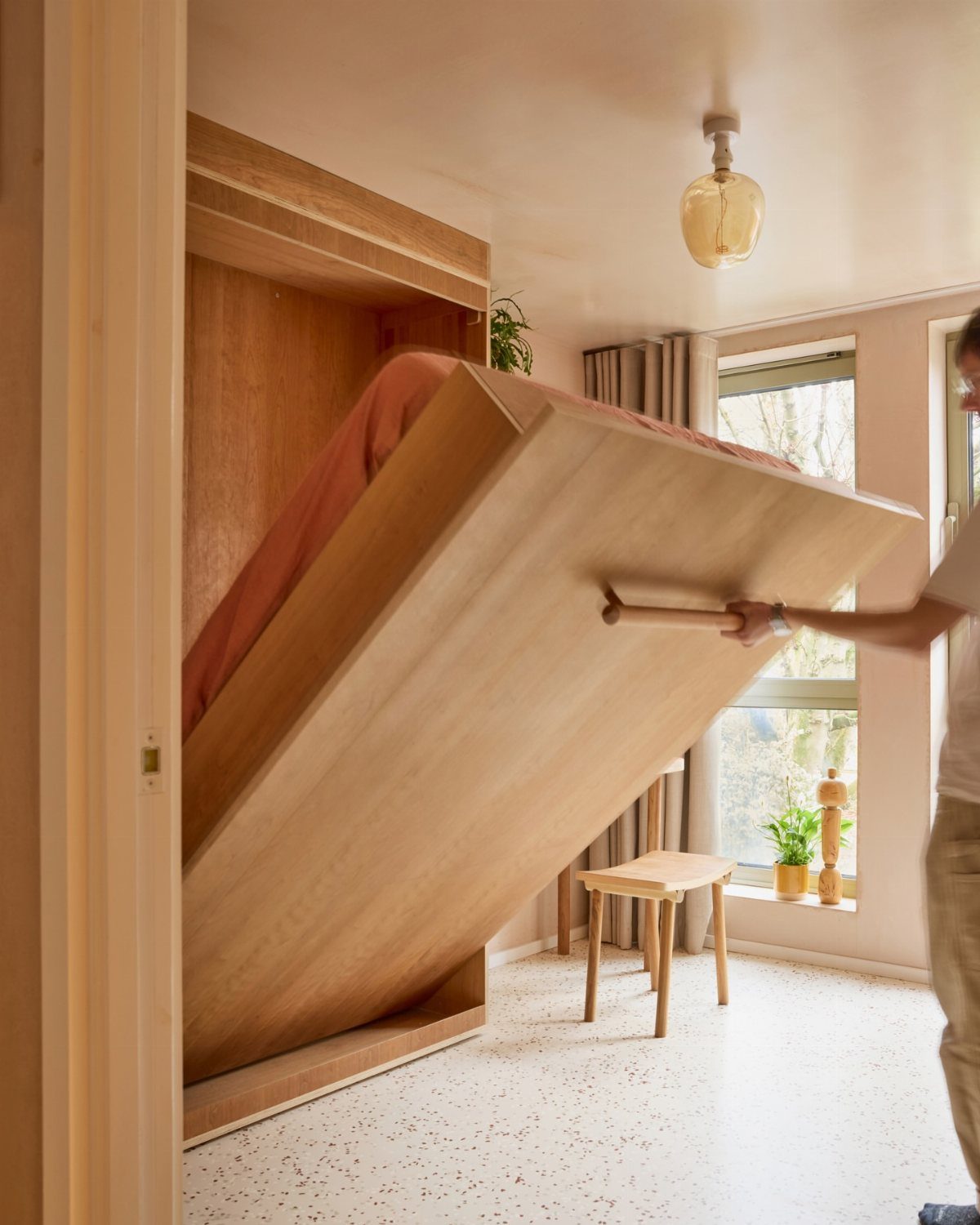 Wooden Murphy bed being lowered from a wall cabinet in a bright room.
