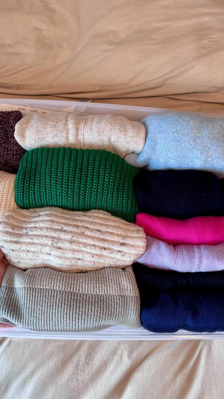 Clear plastic bin filled with folded colorful sweaters on a tan fabric surface.