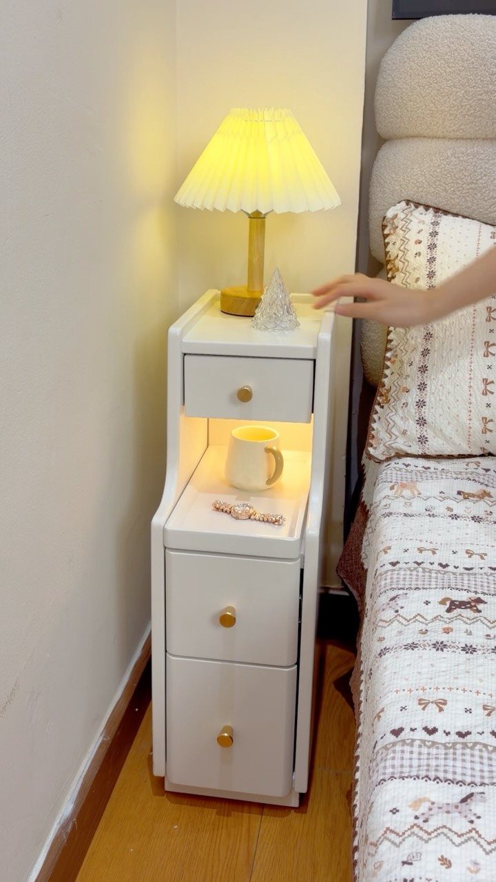 Narrow white bedside table with drawers and a lamp next to a bed.