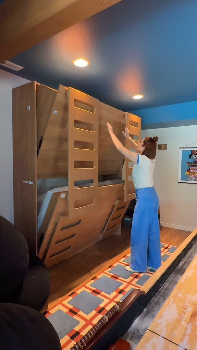 Woman standing next to a light-wood folding bunk bed in a blue room.