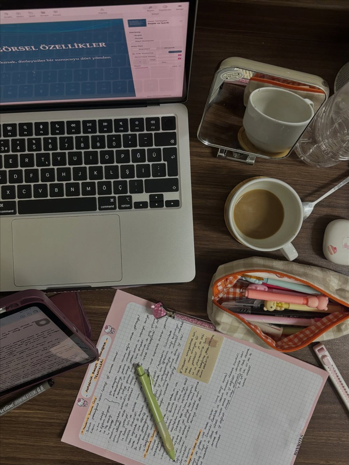 Study desk with laptop, coffee, handwritten notebook, and colorful pens on wood.