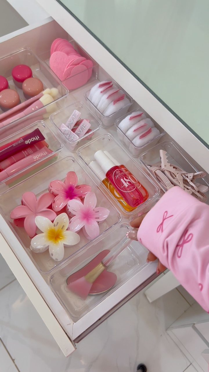 Clear plastic drawer organizers holding pink makeup brushes, skincare, and floral accessories.