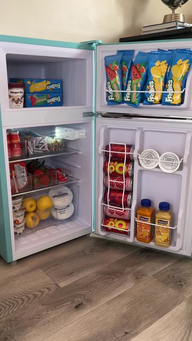 Light-blue mini fridge open and stocked with snacks, drinks, and fresh fruit.