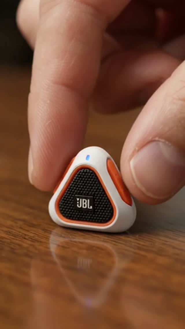 Small triangular JBL wireless speaker held by fingers on a wooden table.