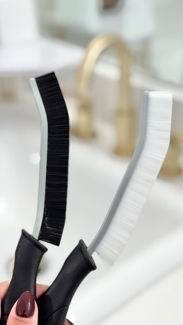 Two narrow cleaning brushes with black and white bristles held near a sink.