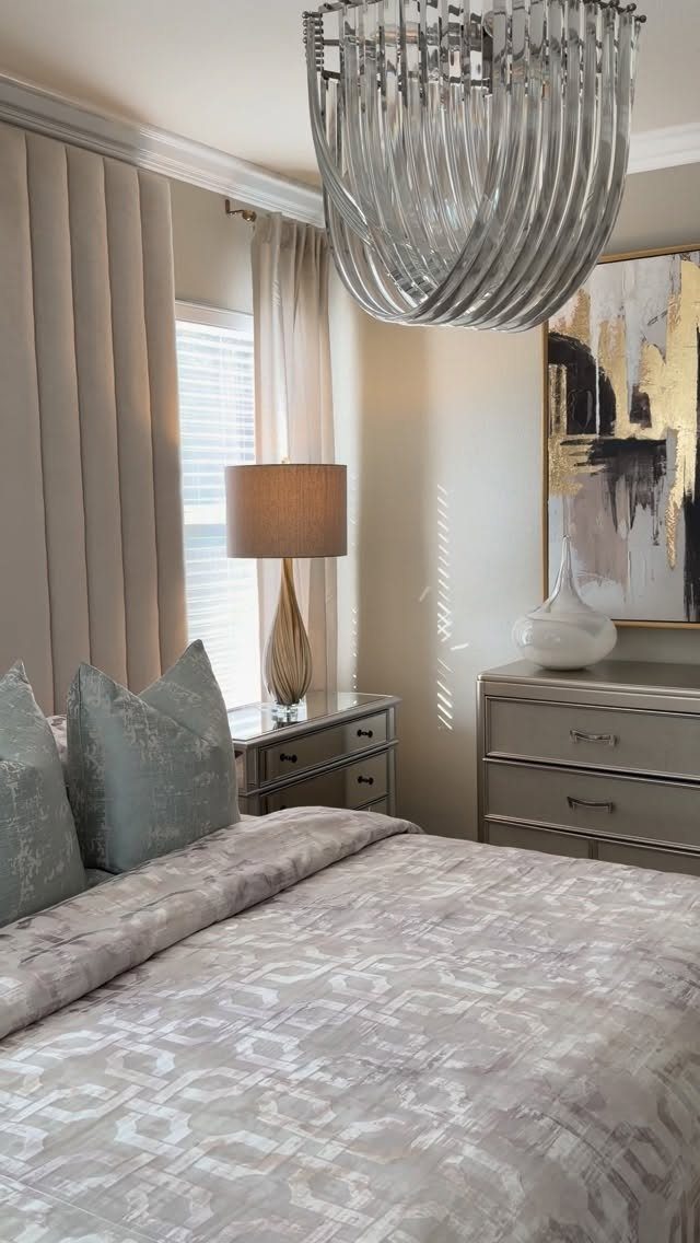 Taupe bedroom walls with a patterned bed, grey dresser, and silver chandelier.