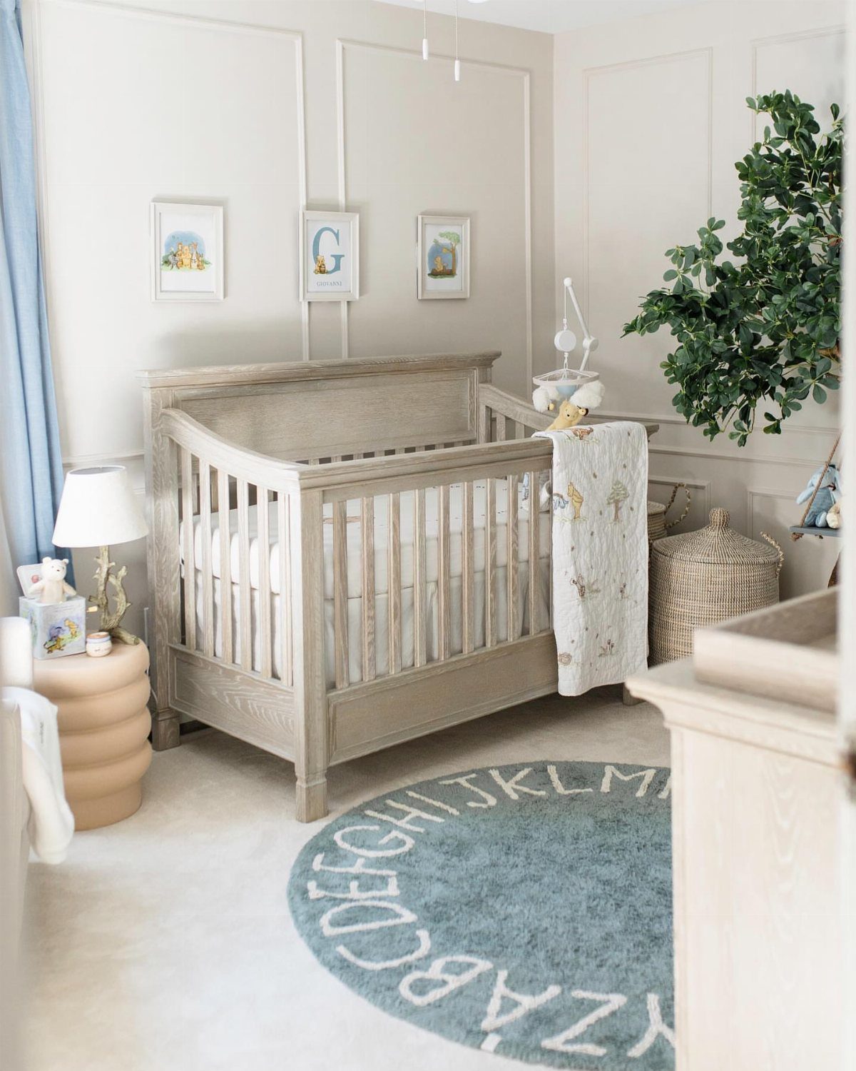 Wooden crib in a neutral nursery with a blue alphabet rug and honey-pot table.